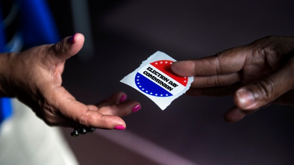 One person giving another person what looks like an "I voted" sticker, but the close-up of the sticker reads "Election Day Communion" instead