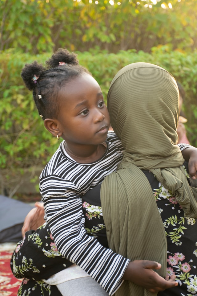 Woman wearing hijab faces away from camera while holding and hugging a young girl, whose open face looks with curiosity past the viewer